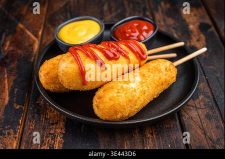 Cani di mais dorato con condimento ketchup serviti su piatto con salsa, snack fast food croccante su sfondo scuro Foto Stock