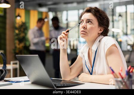 Analista donna caucasica con penna appoggiata su Temple, pensando a strategie finanziarie per la società di marketing. Una donna dirigente lavora sul posto di lavoro, con un profondo pensiero, pianificando un progetto aziendale di successo. Foto Stock