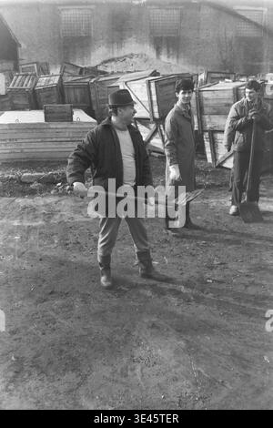 Sloviansk, aprile 1989, cantiere AIZ. Un allegro direttore di fabbrica che indossa un cappello classico si erge orgogliosamente tenendo in mano una pala pesante orizzontalmente come un'arma durante un breve momento di riposo in un tradizionale evento di pulizia volontaria del subbotnik primaverile. Sorride calorosamente mentre è circondato dai suoi colleghi osservatori nel cortile industriale pieno di casse di legno. Questo ritratto sincero riflette meravigliosamente l'umore positivo, l'umorismo e la forte cameratismo del collettivo operaio nel pacifico Donbas. Foto Stock