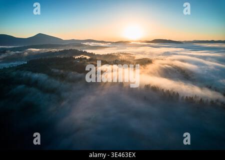 Splendida vista aerea dell'alba sul paesaggio montuoso, con una fitta nebbia che ricopre valli e colline. I raggi dorati del sole penetrano attraverso la nebbia, proiettando una luce serena sull'intera scena. Foto Stock