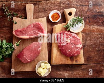 Tagli multipli di costata cruda, roast beef, fianchi e bistecca di roba sono disposti su due assi di legno. Le erbe e le spezie fresche sono collocate in piccole ciotole ne Foto Stock