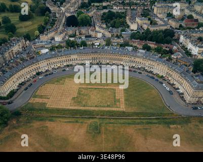 Vista aerea della pietra pallida della Royal Crescent che si snoda elegantemente intorno a un prato soleggiato, fiancheggiato da automobili, un netto contrasto con gli alberi verdi scuri e il Foto Stock