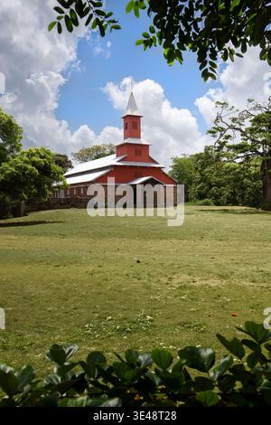 Vista della storica chiesa rossa e bianca con una base in pietra circondata da lussureggianti alberi verdi e un campo erboso sotto un cielo nuvoloso a Iles du Salut, Guy Foto Stock