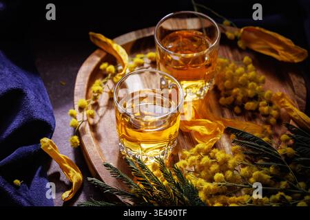 Due bicchieri di whisky ambrato con fiori di mimosa su vassoio in legno concetto di bevanda primaverile Foto Stock