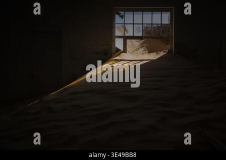 Vista di una camera abbandonata piena di dune di sabbia del deserto e luce del sole che scorre attraverso una finestra rotta a Kolmanskop, regione di Karas, Namibia. Foto Stock