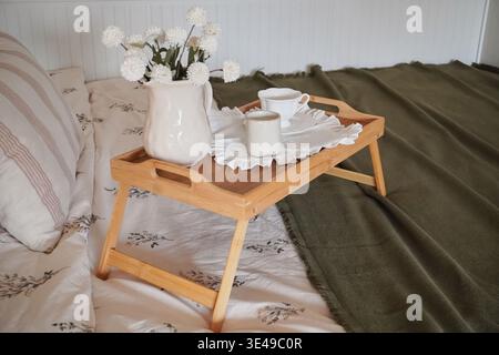 Un accogliente vassoio per la colazione con fiori e bevande su un letto circondato da tessuti morbidi Foto Stock