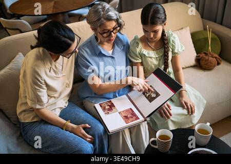 Donna anziana con figlia e ragazza adulta che recensisce album fotografici di famiglia, condividendo i ricordi a casa. Utile per genealogia, eredità familiare, generazioni, marketing professionale, scatti ad angolo alto Foto Stock