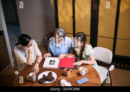 Vista ad angolo alto della donna anziana con figlia e ragazza adulta che recensiscono album fotografici di famiglia a tavola, condividendo ricordi e eredità. Adatto per storia familiare, generazioni, marketing generazionale Foto Stock