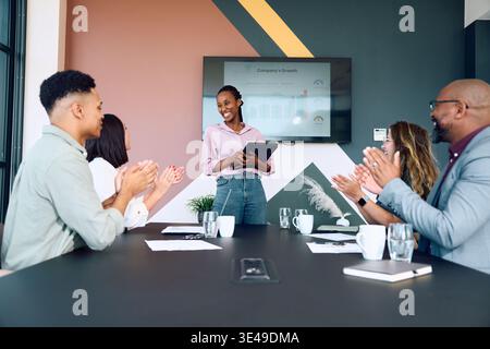 Giovane donna d'affari sicura che tiene in mano un tablet e sorride mentre il team applaude in sala riunioni in ufficio Foto Stock