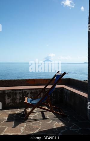 Vista di una sedia a sdraio blu su una terrazza in pietra che si affaccia sul Mar Tirreno con il vulcano Stromboli fumante all'orizzonte sotto un cielo limpido a Sal Foto Stock