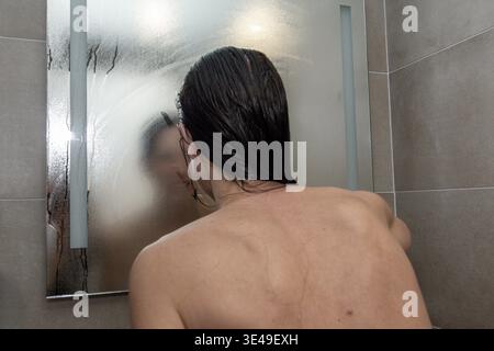 La donna si trova in bagno e pulisce con la mano uno specchio a vapore dopo una doccia, i capelli bagnati e la luce soffusa del mattino, suggerendo una cura di sé tranquilla e intima Foto Stock