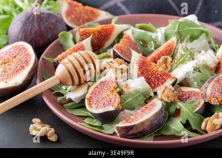 Gustosa insalata con fichi a fette, formaggio di capra soffice tritato, noci e foglie di rucola fresca con cucchiaio di miele in legno su piatto di ceramica rosso retrò su br scuro Foto Stock