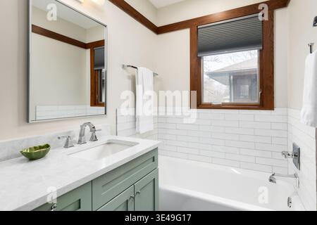 Un lavandino del bagno con rubinetto cromato su un ripiano in marmo bianco, armadio verde salvia e vasca rivestita con piastrelle metropolitane in ceramica bianca. Foto Stock
