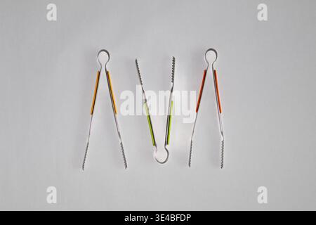 Composizione piatta minima delle pinze da cucina in acciaio inox con manici colorati in silicone su sfondo bianco. Utensili da cucina moderni Foto Stock