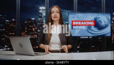 Anchor donna sicura di sé che presenta notizie, guarda la fotocamera e parla alla White Desk. Concetto di giornalismo televisivo high-tech con laptop, schermo digitale e vista notturna sfocata della città. Verticale. Foto Stock