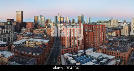 Un ampio panorama aereo che cattura lo skyline di Manchester mentre il sole nascente proietta un bagliore dorato su un mix di mattoni storici e moderne torri di vetro. Foto Stock