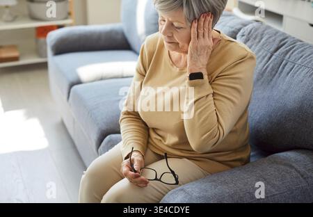 Triste donna anziana stanca con mal di testa seduta sul divano a casa, soffre di dolore Foto Stock