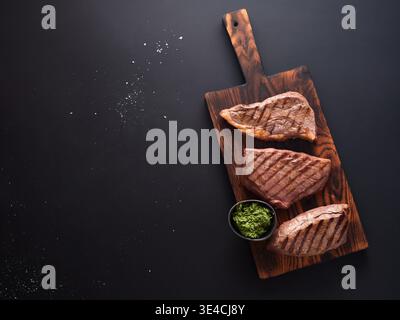 Il fianco di roast beef alla griglia e la bistecca di roast sono esposti su un asse di legno. Un piccolo recipiente di salsa verde è collocato nelle vicinanze. La scena mostra un tavolo da cucina Foto Stock