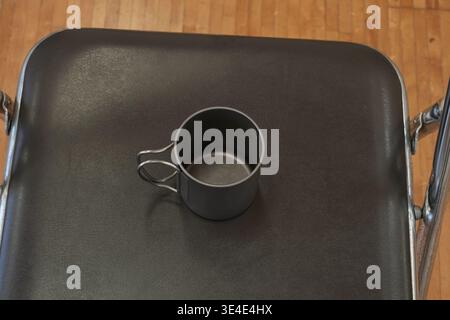 La tazza scura poggia su una superficie nera ruvida. Scena semplice e minimalista con linee pulite. Il manico metallico si curva leggermente di lato. Design robusto e funzionale Foto Stock