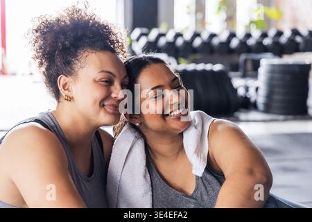 Amiche afroamericane che sorridono, si appoggiano e indossano canotte in palestra con manubri e asciugamano Foto Stock