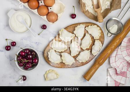 Cibo, prepara deliziosi gnocchi dolci con ciliegie. Dessert tradizionale russo e ucraino. Vareniki e ingredienti su fondo in marmo -f Foto Stock