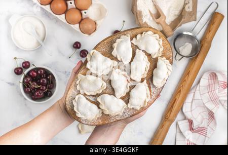 Cibo, prepara deliziosi gnocchi dolci con ciliegie. Dessert tradizionale russo e ucraino. Vareniki e ingredienti su fondo in marmo -f Foto Stock