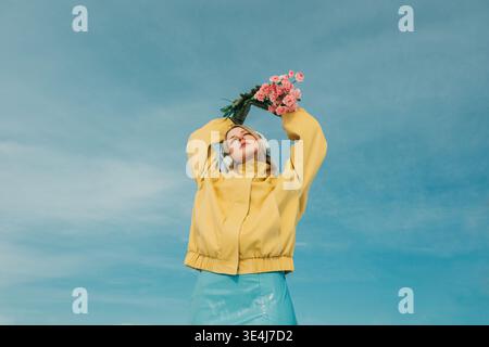 Una donna con una giacca gialla e una gonna blu si staglia all'aperto contro un cielo blu brillante, indossando le cuffie e tenendo i fiori rosa, esprimendo gioia e Foto Stock