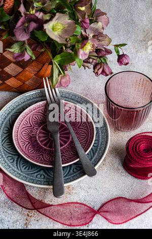 Elegante tavolino primaverile con bouquet a forma di alce, piatti in ceramica a motivi geometrici, posate scure e bicchieri viola su una superficie in pietra con r rosso Foto Stock