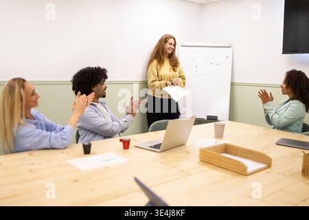 Un team diversificato applaude un collega che presenta idee in una luminosa sala riunioni dell'ufficio di coworking, con laptop e appunti sul tavolo, mettendo in risalto il collaboratore Foto Stock