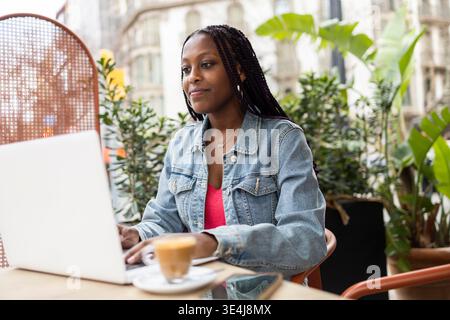 Una donna lavora con il proprio laptop in un accogliente caffè all'aperto nel quartiere Gracia di Barcellona, Spagna, catturando l'essenza di uno stile di vita moderno e rilassato Foto Stock