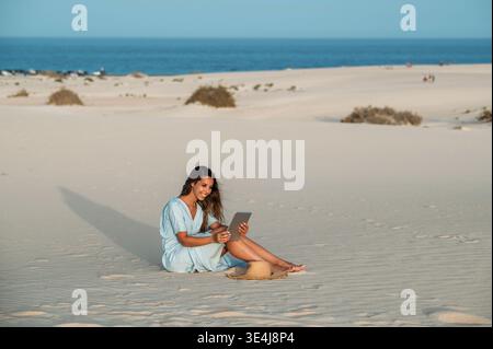 Una giovane turista siede sulle dune di sabbia a Corralejo, usando un tablet in una giornata di sole a Fuerteventura, Isole Canarie, Spagna, godendosi rilassato Foto Stock