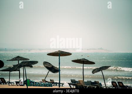 Vista tranquilla della spiaggia di Essaouira in Marocco, con ombrelloni di paglia e lettini vuoti sull'Atlantico, con onde dolci e cielo nebbioso che creano una calma Foto Stock