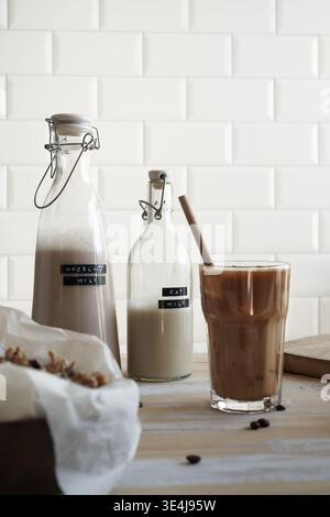 Bicchiere alto di caffè ghiacciato mescolato con latte e cannuccia di legno, con due biberon ribaltabili etichettati LATTE DI NOCCIOLE e LATTE di avena, parzialmente riempiti Foto Stock
