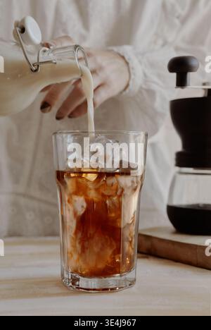 Primo piano di mani che versano il latte in un bicchiere di caffè ghiacciato con cubetti di ghiaccio, creando un turbinio cremoso su un bancone luminoso accanto a una caraffa fredda in un c Foto Stock