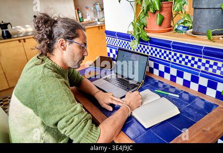 Un ispanico lavora da casa al bancone della cucina. Sta scrivendo appunti e usando un portatile. La cucina è ordinata con piante e utensili visibili. IT Foto Stock