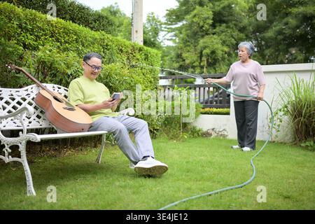 Una coppia anziana si diverte a fare giardinaggio insieme nel loro giardino di casa. Una donna che innaffia le piante con un tubo mentre suo marito si rilassa su una panchina utilizzando uno smartphone, mostrando uno stile di vita pensionistico felice || modello rilasciato Foto Stock