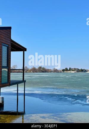 Una foto verticale di un'accogliente terrazza in legno su palafitte che si affaccia su un lago parzialmente ghiacciato con ghiaccio turchese. Sullo sfondo, lussuosa vista lago vi Foto Stock