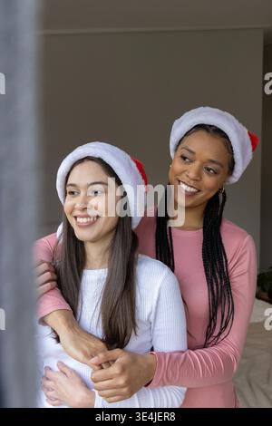 Diverse partner femminili in piedi e abbracciati in camera da letto, indossando cappelli Babbo Natale e comodi top. Natale, copricapo, calore, intimo, coppia, Home, illuminazione || modello rilasciato Foto Stock