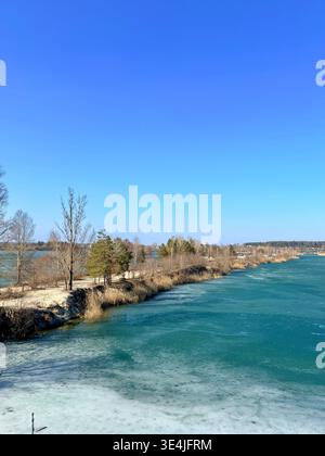Una vista panoramica ad alto angolo di uno splendido lago turchese ghiacciato diviso da una sottile penisola sabbiosa con alberi sparsi. La scena viene catturata sotto un cri Foto Stock