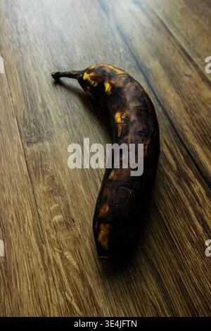 Banana troppo matura con macchie scure sulla superficie in legno, che illustra sprechi di cibo, deterioramento e decadimento naturale in una scena di natura morta minima Foto Stock