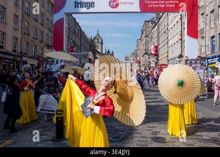 Artisti di strada colorati in costumi tradizionali gialli e rossi ballano con ombrelloni di carta in una trafficata strada di Edimburgo durante il festival Fringe Foto Stock