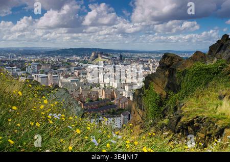 Vista panoramica da Salisbury Crags sullo skyline di Edimburgo, fiori selvatici in primo piano e scogliere rocciose con lo storico Castello di Edimburgo sotto il suggestivo c Foto Stock