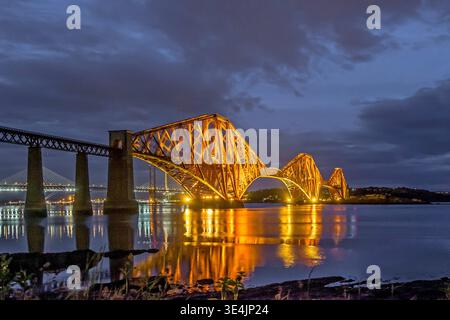 Il Forth Rail Bridge si illumina d'oro contro un cielo al crepuscolo, mentre il suo sbalzo in acciaio si estende nella calma Firth of Forth Waters, mettendo in risalto la terra Foto Stock