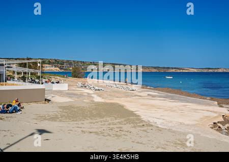 Mellieha, Malta - 6 febbraio 2026: L'ampia baia di Ghadira, sabbiosa, con un caffè sulla spiaggia, lettini e un complesso turistico distante Foto Stock