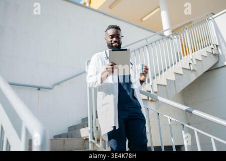 Felice nero maschio medico utilizzando tablet digitale di fronte al moderno edificio ospedaliero Foto Stock