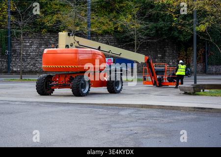 Il sollevatore con braccio arancione parcheggiato su una strada, mentre due operai edili in attrezzature di sicurezza lavorano vicino alla piattaforma, con alberi e pareti di pietra dietro. Foto Stock