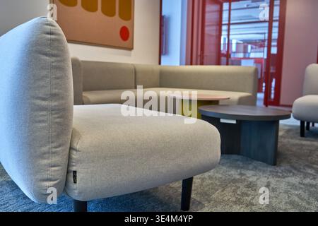 L'immagine raffigura un moderno salotto di ufficio o un interno di un'area d'attesa. In primo piano, una poltrona imbottita grigio chiaro con gambe in legno scuro è Foto Stock