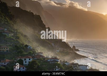 Spettacolare vista aerea della costa di Madeira al tramonto, con vivaci arance e blu. L'oceano sereno, le montagne lussureggianti e l'incantevole villaggio Foto Stock