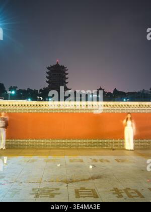 Wuhan, Cina - 17 maggio 2025: Foto a lunga esposizione che mostra figure sfocate che camminano davanti a un lungo muro rosso di notte. L'immagine acquisisce un'attività commerciale illuminata Foto Stock