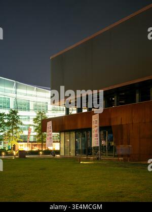 Wuhan, Cina - 11 giugno 2025: Prato erboso di fronte a un moderno edificio marrone e una struttura in vetro illuminata di notte. L'immagine mostra l'esterno Foto Stock